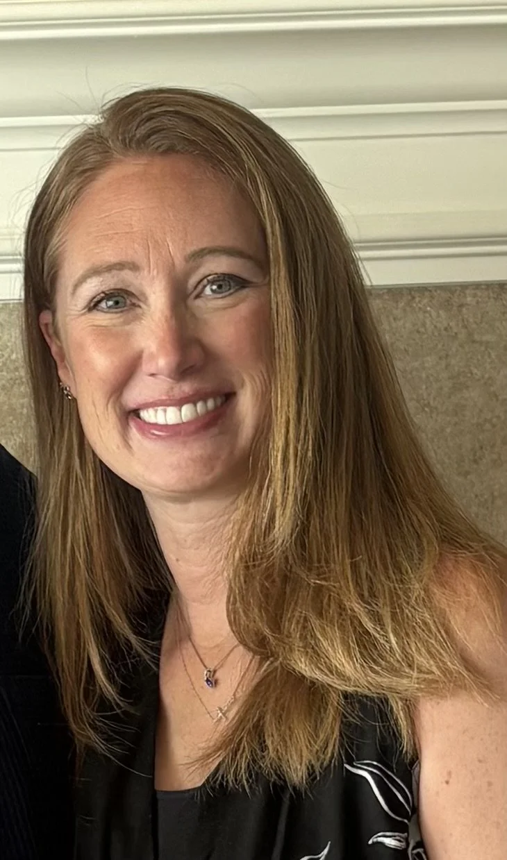 A woman with long, reddish-brown hair and blue eyes smiling at the camera, wearing a black top with a necklace, in front of a light-colored wall with white trim.