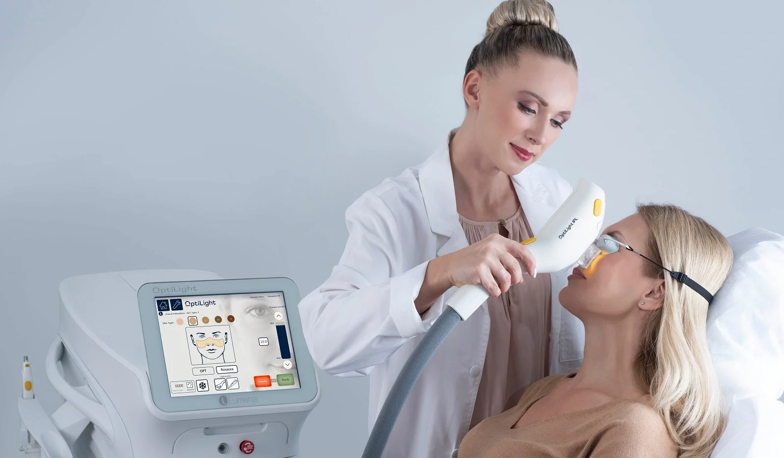 A woman lying on a medical bed receiving a skin treatment from a healthcare professional using a handheld medical device in a clinical setting.