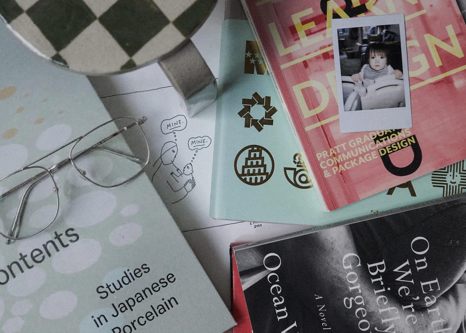 Desk with reading materials including a book titled 'On Earth We Wore Orange', a pamphlet on Japanese porcelain, a semi-rimless eyeglasses, a quarterly report, and a pink pamphlet. There is also a small hand-drawn illustration of a person holding a child with speech bubbles saying 'MINE'.