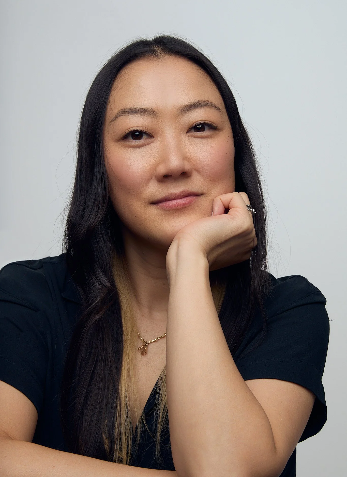 Portrait of a woman with long dark hair, resting her chin on her hand, wearing a black top and jewelry, against a plain light background.