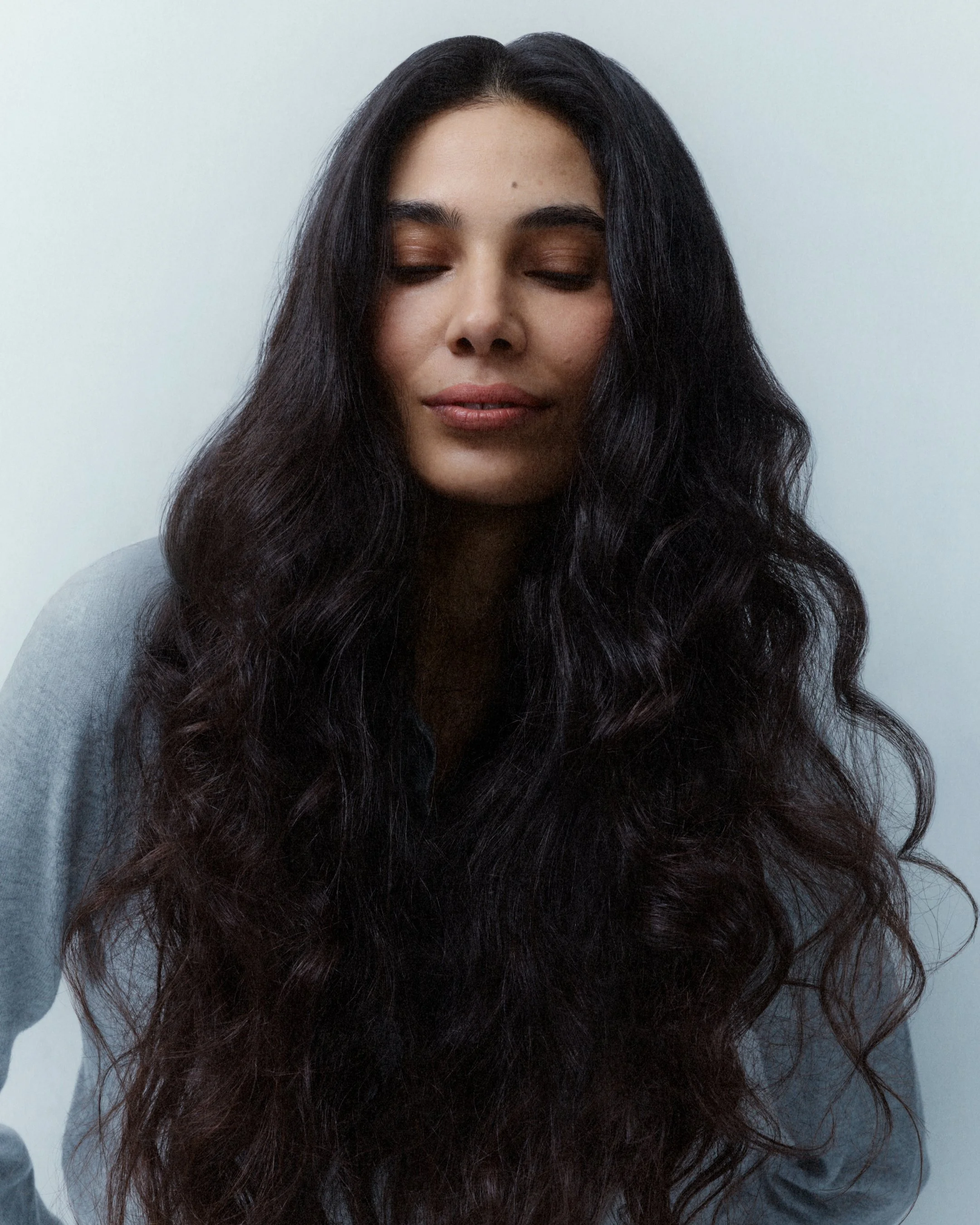 Close-up of a woman with long, dark, wavy hair and her eyes closed, wearing a gray top against a plain light-colored background.