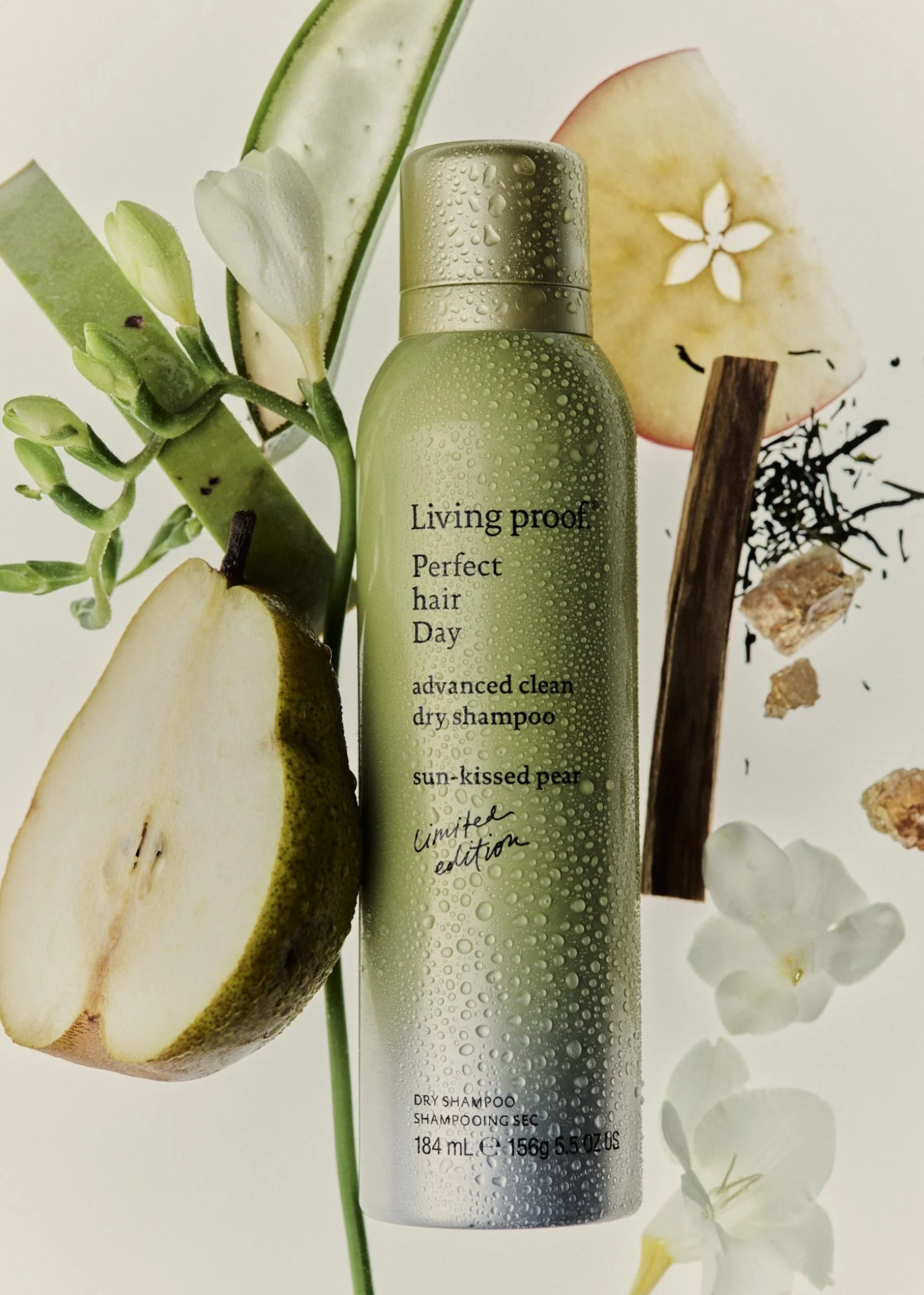 A green bottle of dry shampoo surrounded by sliced pear, cucumber, vanilla bean, dried herbs, and white flowers, with condensation droplets on the bottle.