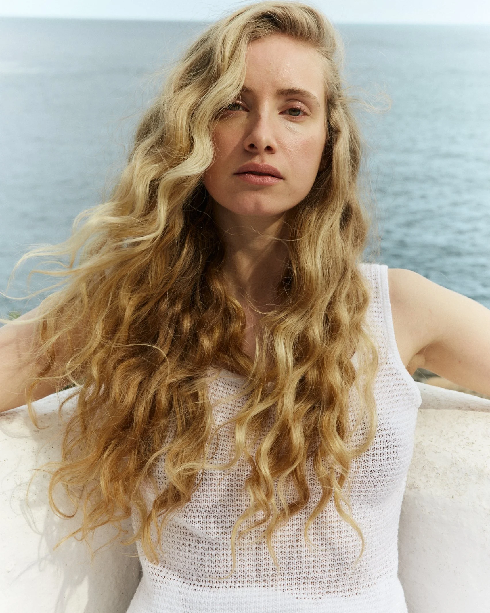 Close-up of a woman with long, wavy blonde hair, wearing a white knitted top, standing outdoors near the water.