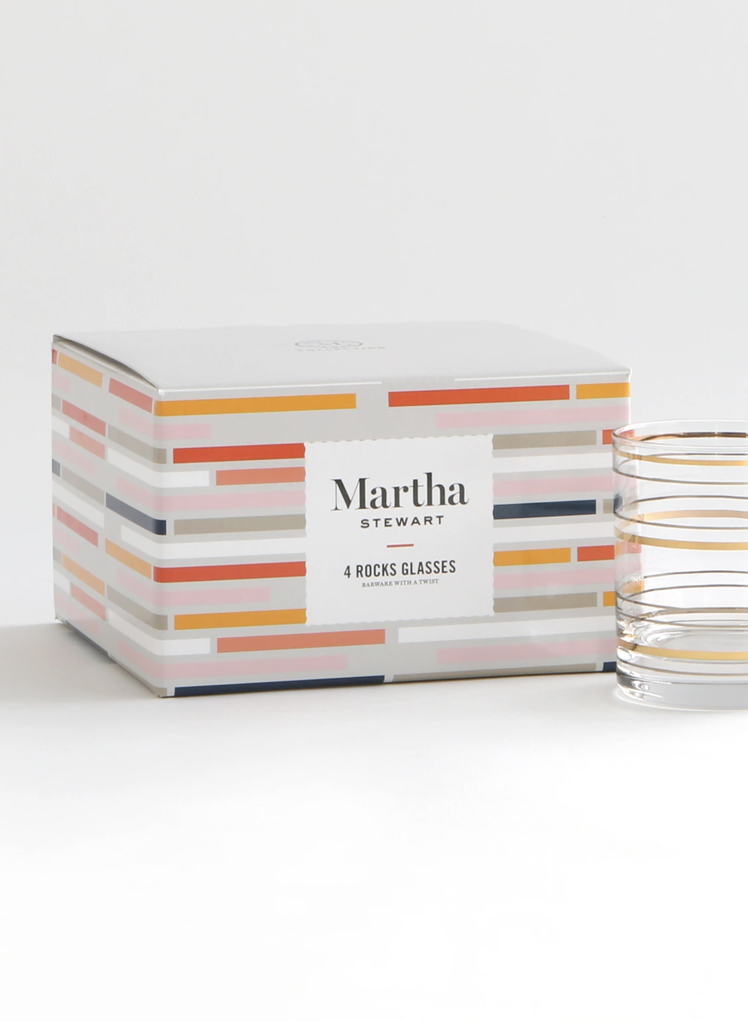 A box of Martha Stewart 4 rocks glasses with colorful striped design, placed on a white surface next to two transparent glasses with gold rims.