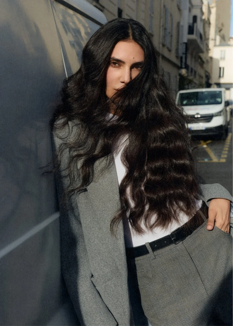 A woman with long, wavy dark hair leaning against a wall on a city street, wearing a gray blazer and plaid pants, with her hand in her pocket.