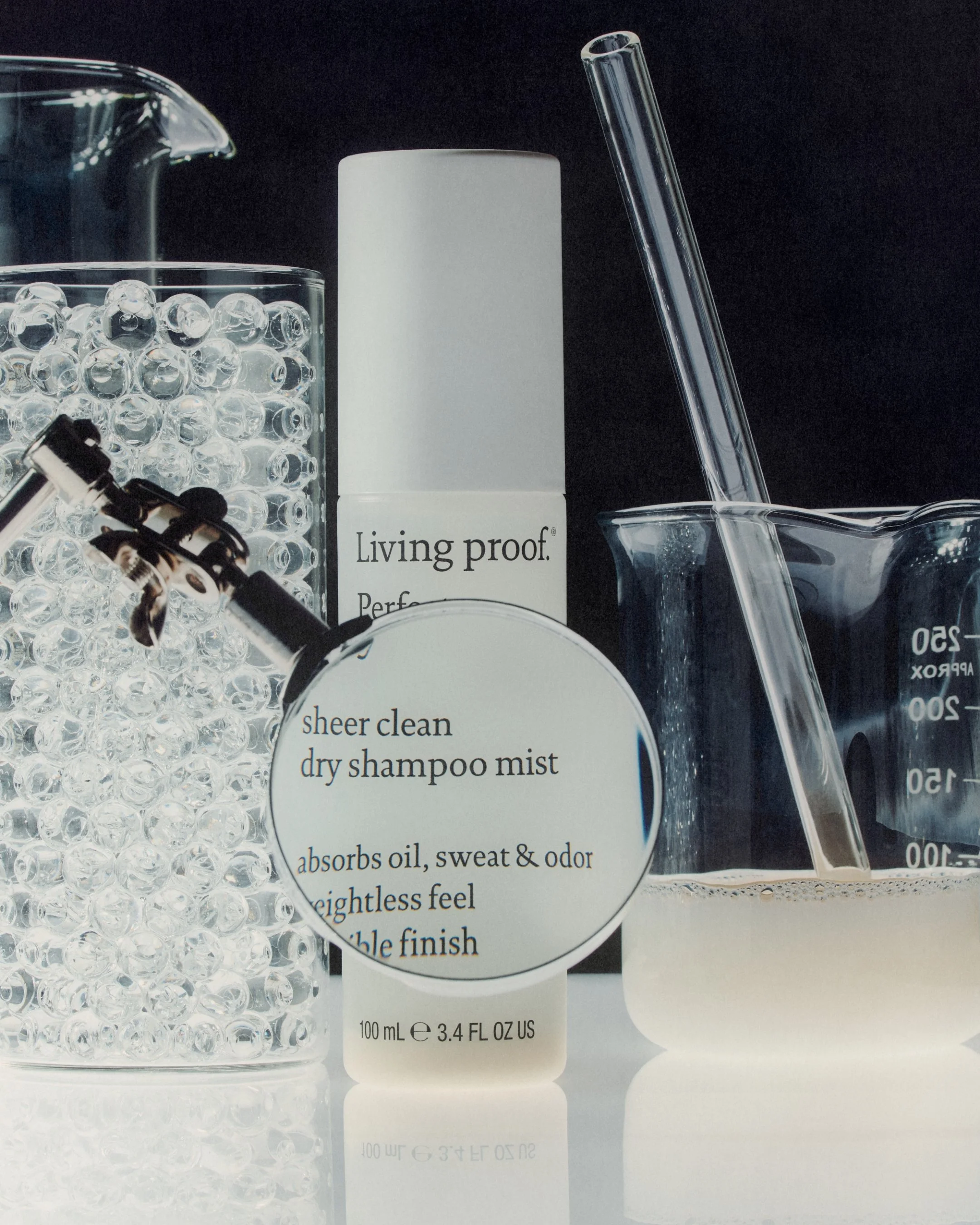 Collection of clear plastic laboratory items, including glass bead container, white bottle labeled 'living proof,' glass dropper, and graduated measuring cup, all against a black background.