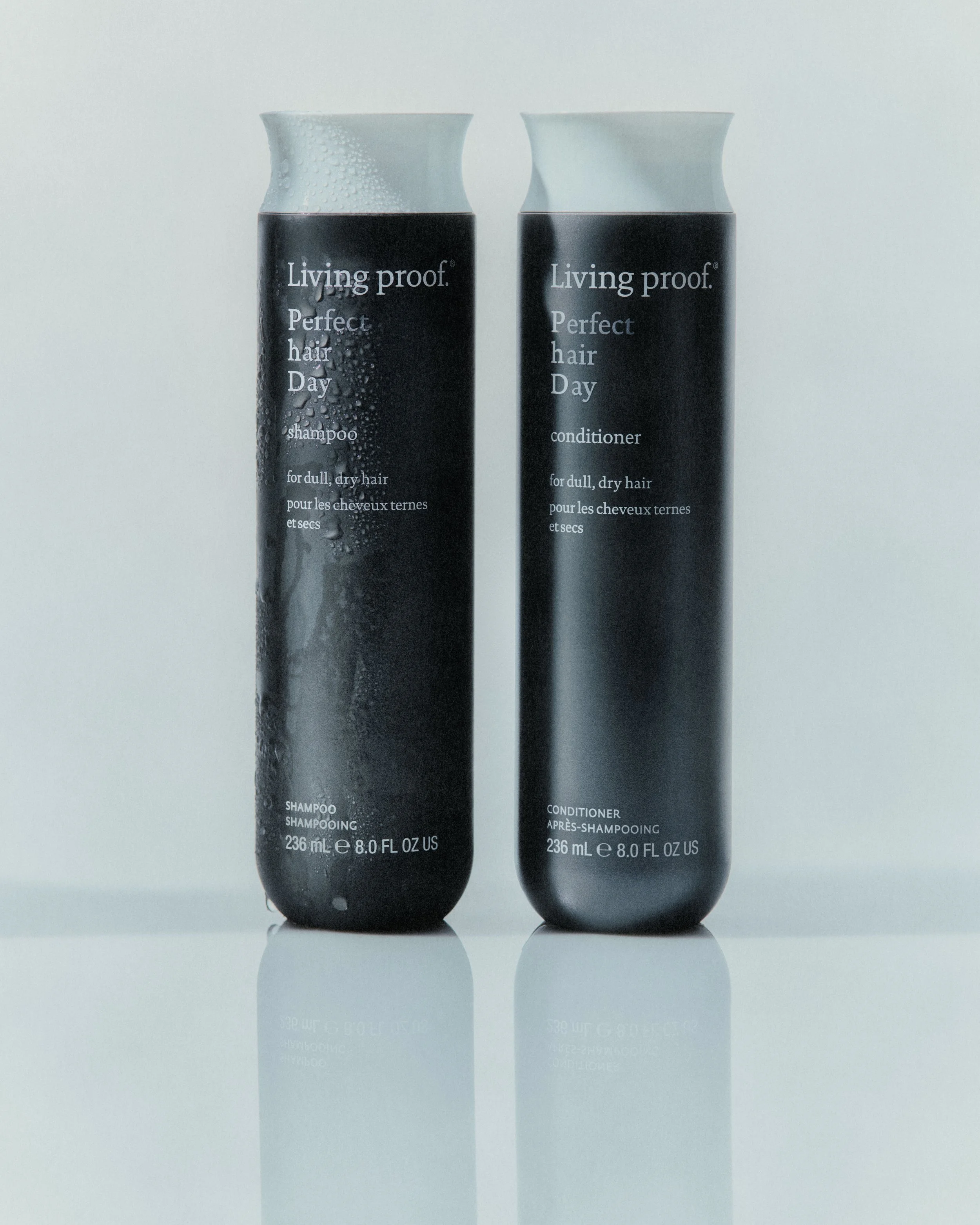 Two black bottles of Living Proof Perfect Hair Day shampoo and conditioner, with white caps, on a reflective surface against a light background.