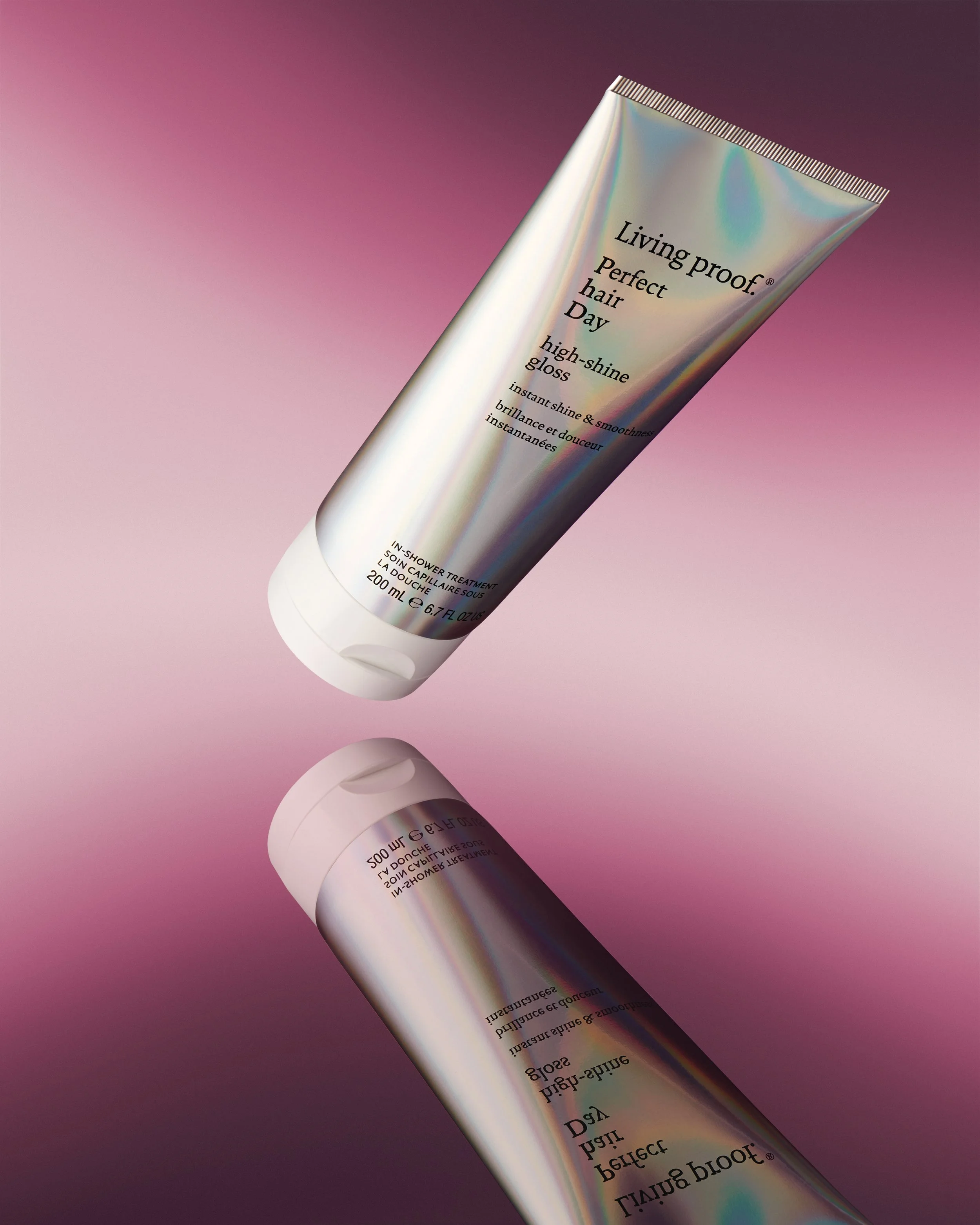 A iridescent tube of Living Proof Perfect Hair Day high-shine gloss with a reflective, rainbow-like surface, resting on a pink gradient background. The tube's label includes product details and branding.