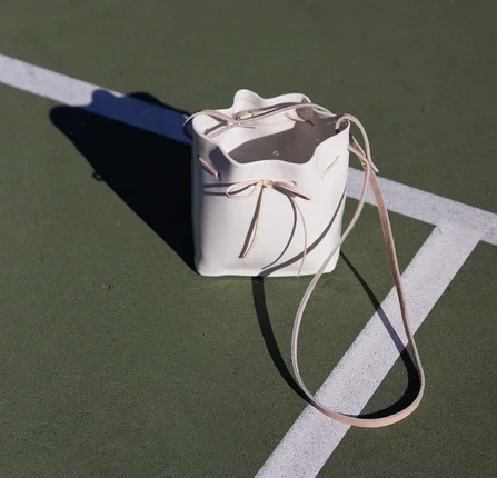 A white handbag with a pink strap resting on a tennis court, casting a shadow.