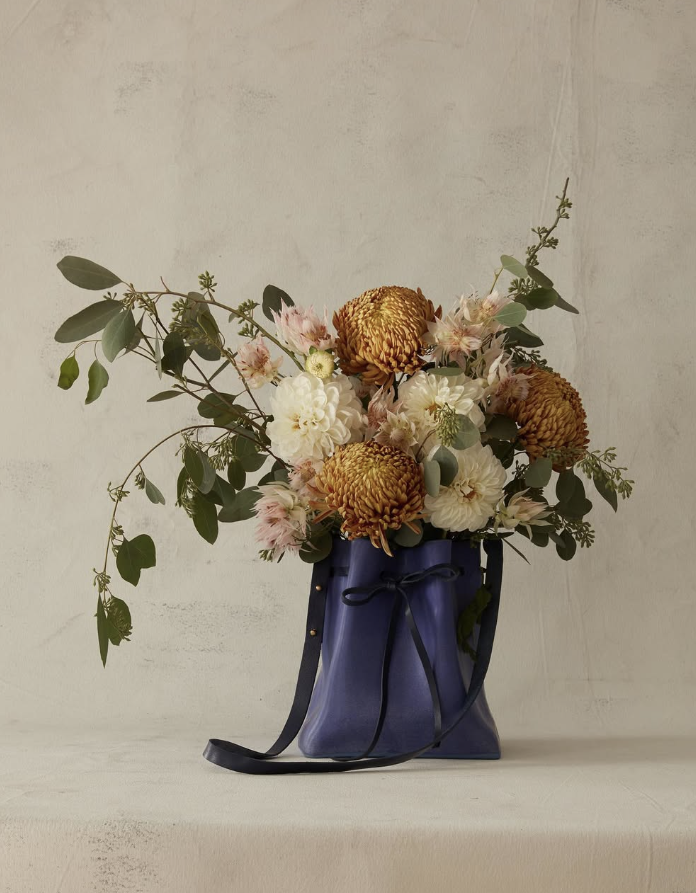 A bouquet of flowers in a dark purple ceramic vase with a black ribbon. The bouquet includes white and pink flowers, large orange flowers, and green foliage. The vase is placed on a plain, light-colored surface against a light-colored wall.