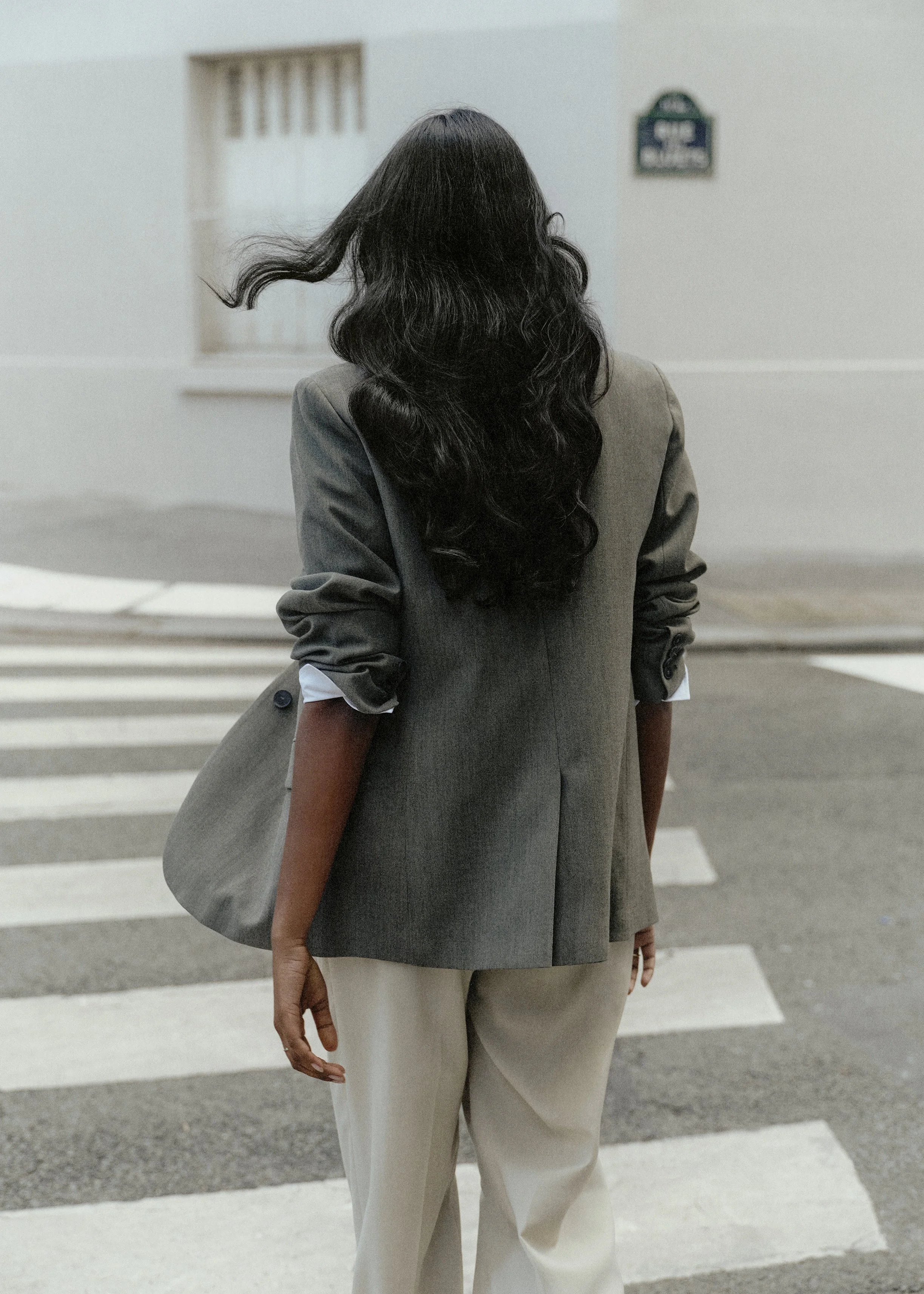 A person with long black hair, wearing a gray blazer and beige pants, is walking across a crosswalk. Their right arm is visible, hanging down with fingers relaxed.