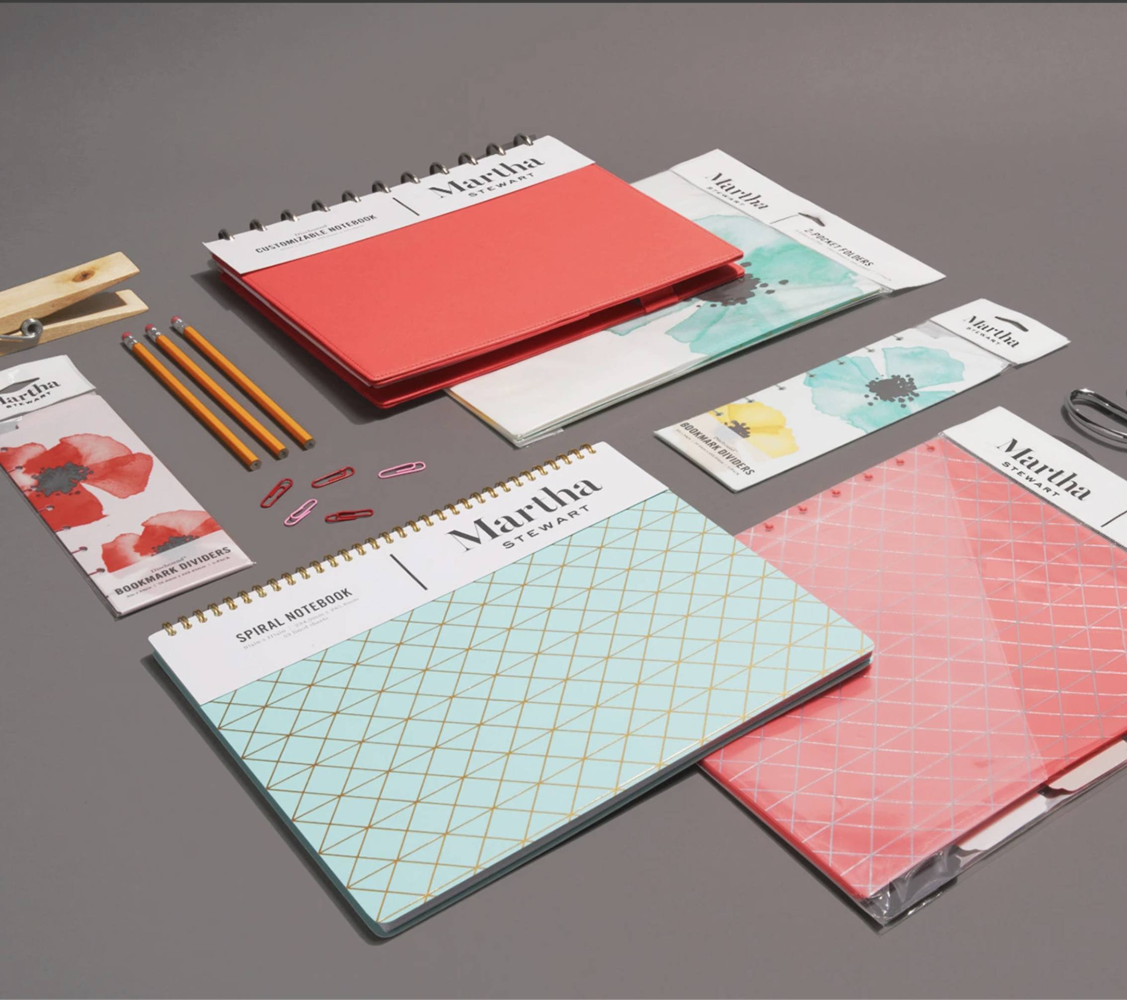 A gray table displays organization and office supplies, including a red binder, a spiral notebook with a geometric pattern cover, and various bookmarks and paper clips. The items are neatly arranged for planning or creative work.