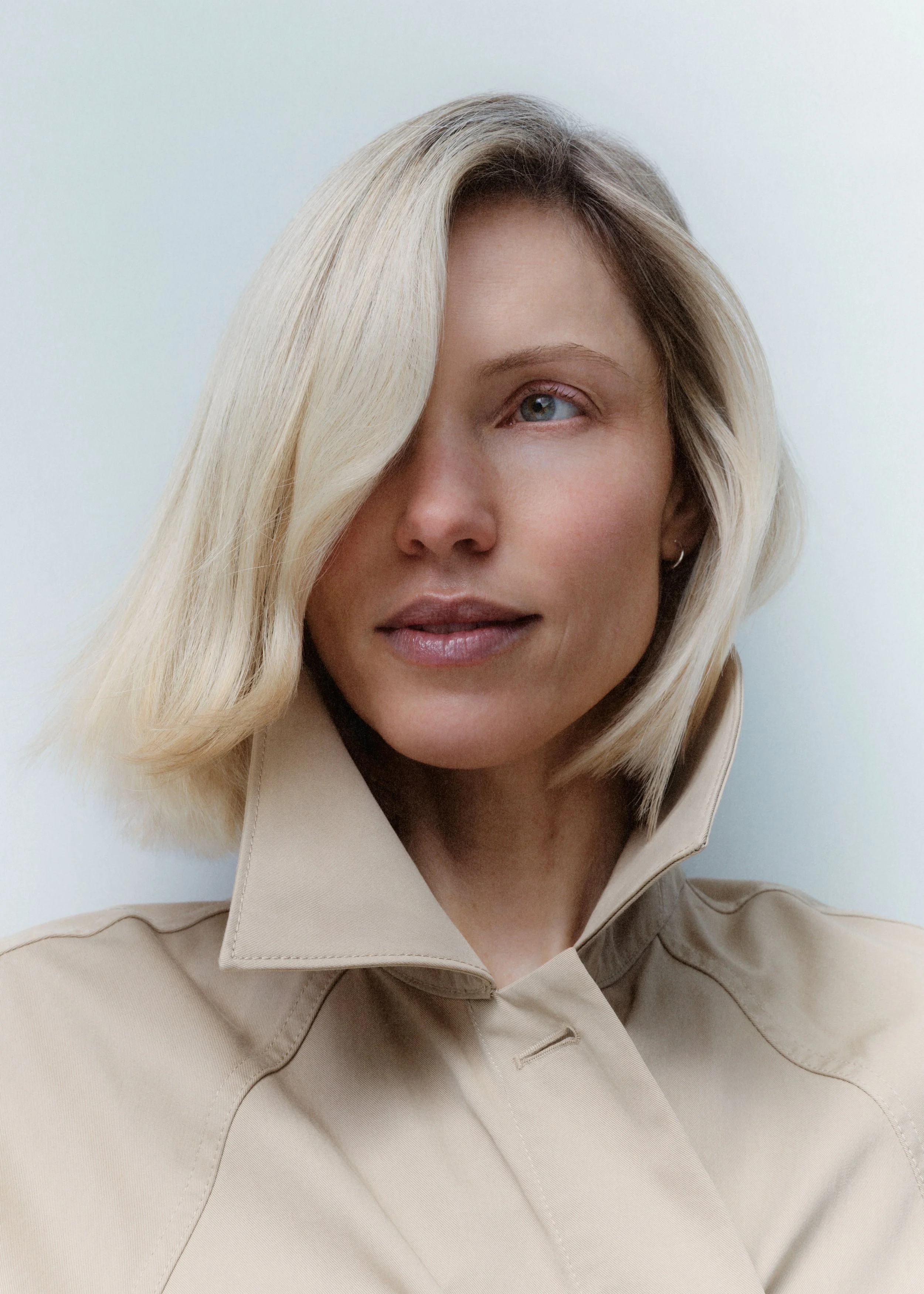 Close-up portrait of a woman with blonde hair and blue eyes, wearing a beige collared shirt, looking slightly to the side, against a plain light background.
