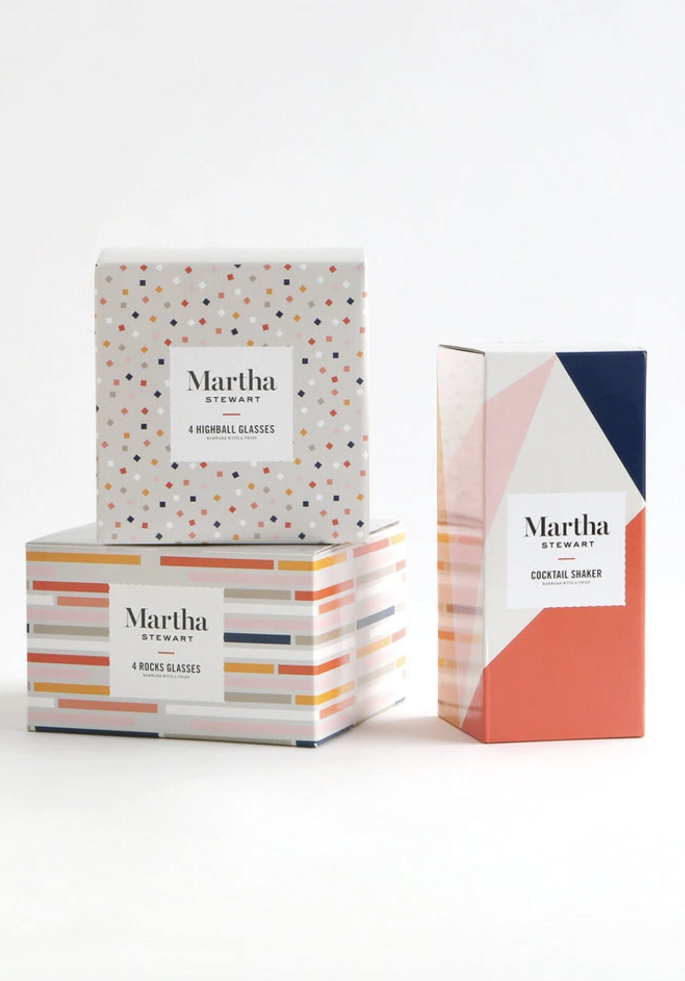 Three colorful boxes of Martha Stewart glassware, including highball glasses, rocks glasses, and a cocktail shaker, arranged on a white surface against a plain white background.