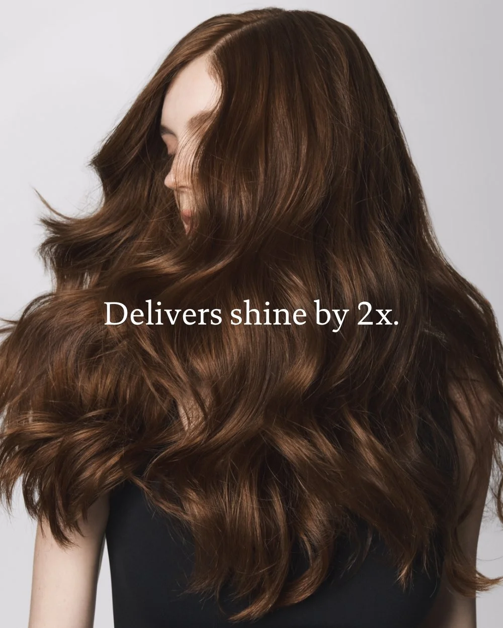 Close-up of a woman with long, wavy, shiny brown hair, partially covering her face, with a plain light gray background. Text overlay says 'Delivers shine by 2x.'
