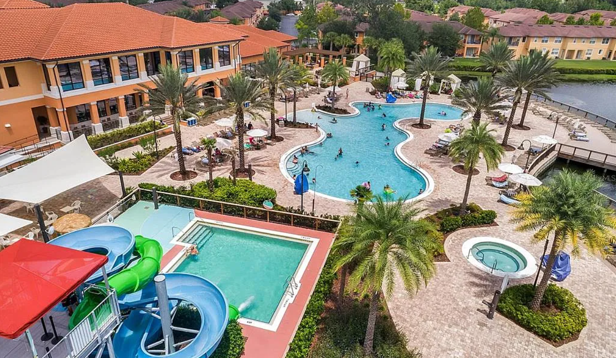 Aerial view of a resort-style pool area with water slides, hot tub, palm trees, lounge chairs, umbrellas, and people swimming and relaxing.