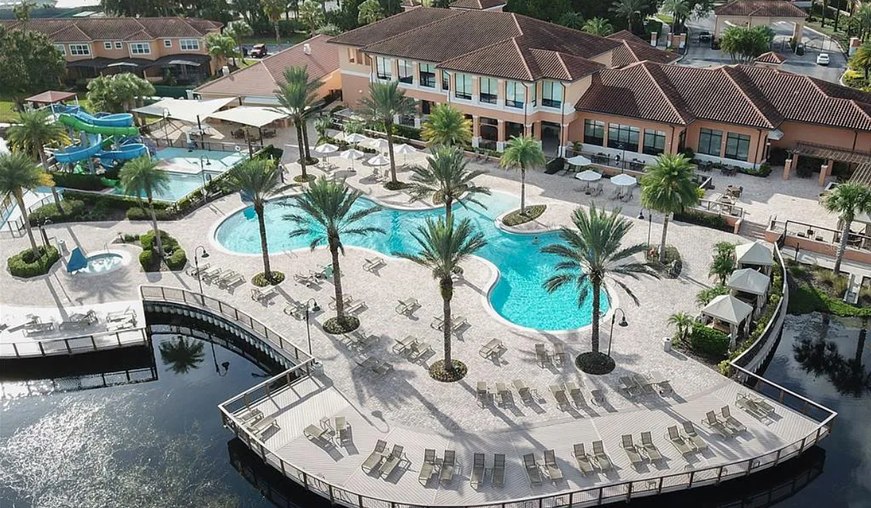An aerial view of a resort pool area with palm trees, lounge chairs, and water slides, surrounded by buildings and a pond.
