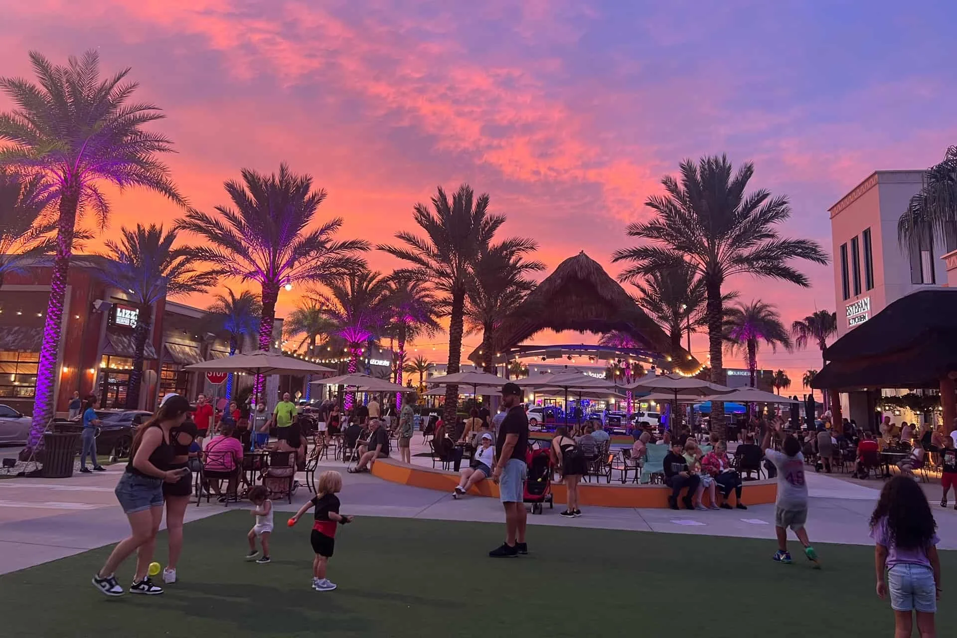 Promenade at Sunset Walk outdoor entertainment area with palm trees, colorful lights, and visitors at sunset.