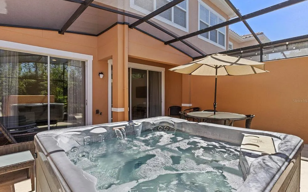 An outdoor patio area with a hot tub, patio table with an umbrella, chairs, and a screened enclosure, attached to a multi-story house.