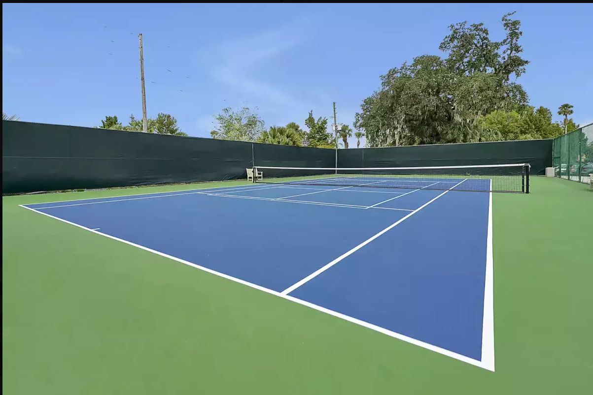 Empty outdoor tennis court with blue playing surface and white lines, surrounded by a green fence, with trees and a blue sky in the background.