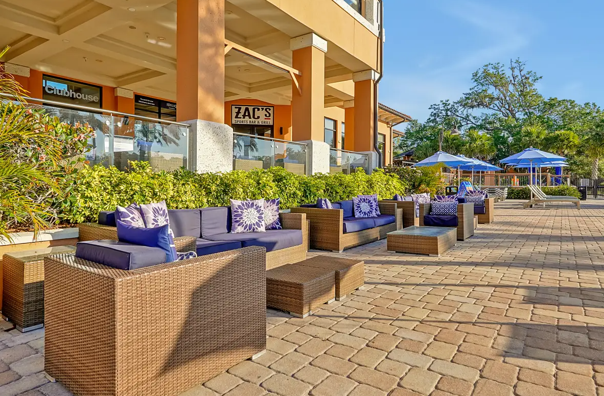 Outdoor patio area with wicker sofas and purple cushions, surrounded by green bushes and trees, with umbrellas and lounge chairs in the background, near a building with signs for a sports bar and a nightclub.