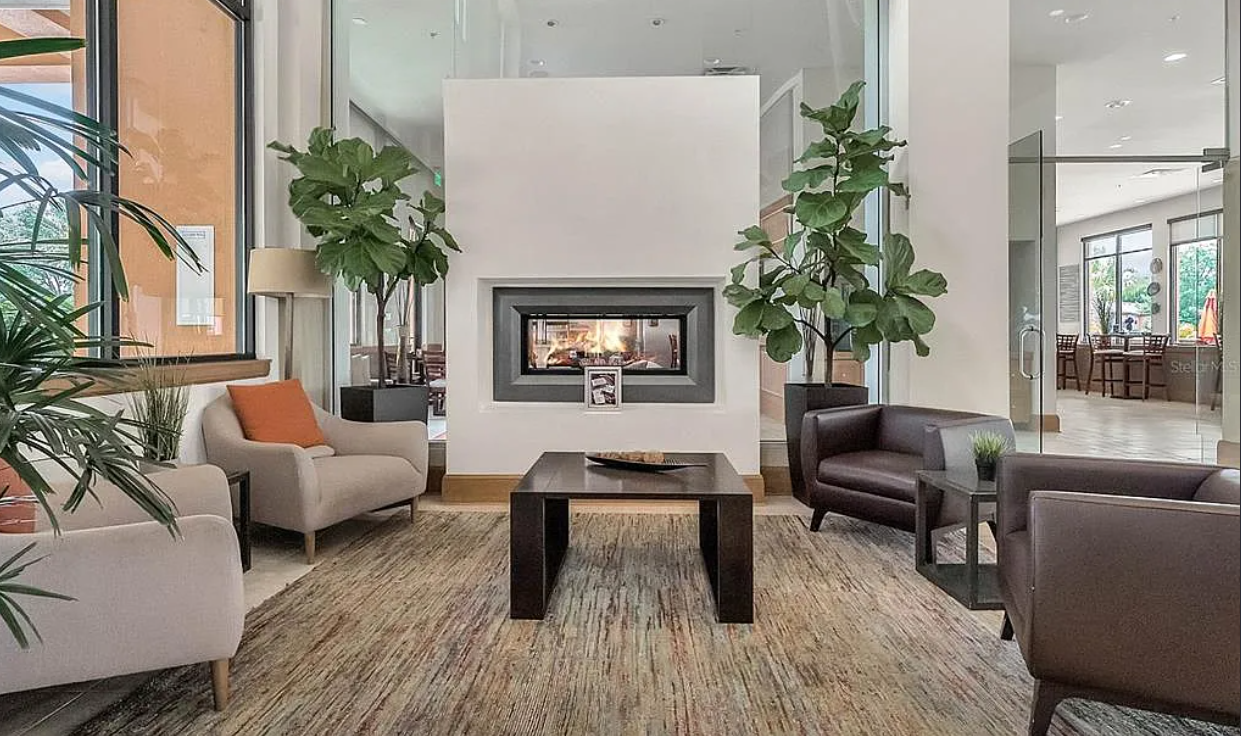Modern lobby with fireplace, beige and dark brown chairs, potted plants, and a wooden coffee table on a textured rug.