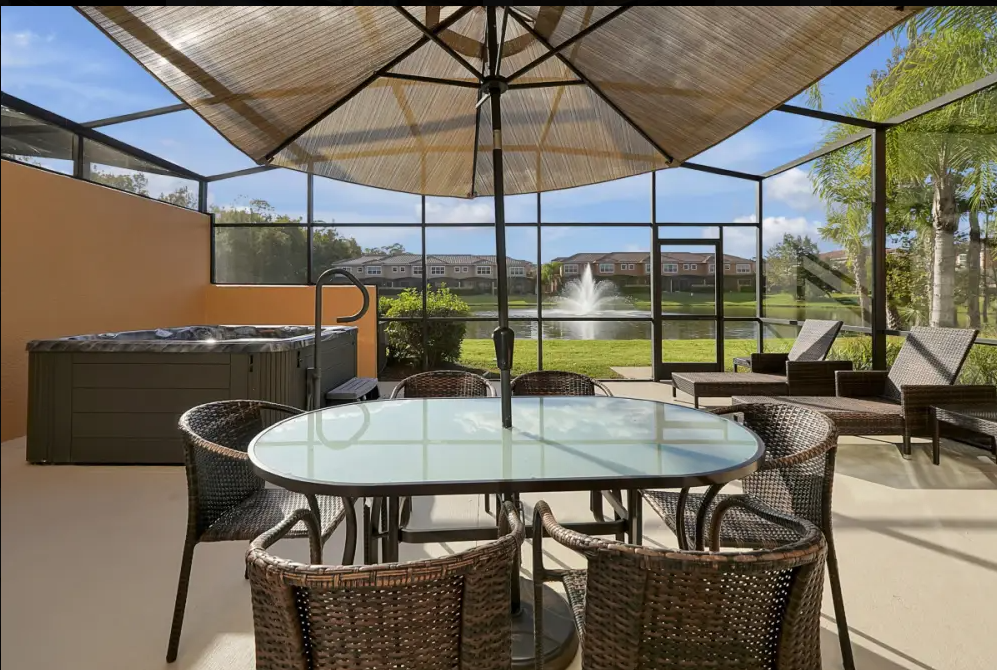 Enclosed patio with a large glass-top dining table, six wicker chairs, a hot tub, and two lounge chairs overlooking a pond with a fountain, greenery, trees, and residential buildings in the background.