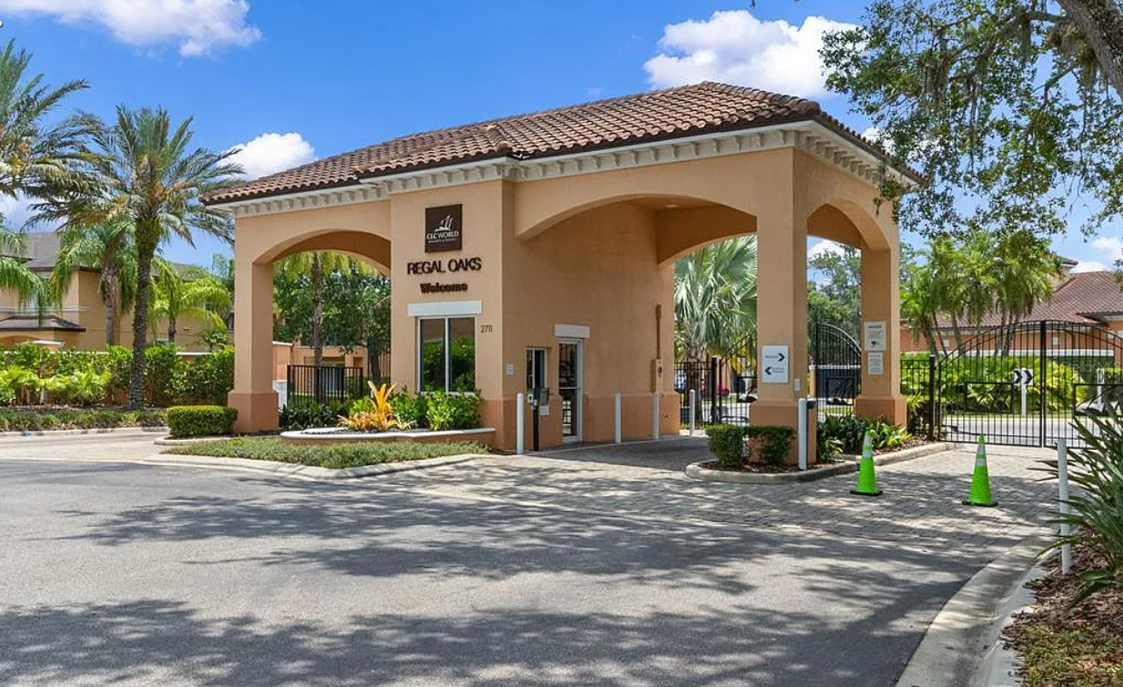 Entrance to a gated residential community called Regal Oaks with palm trees, landscaping, and a small guardhouse or security booth.