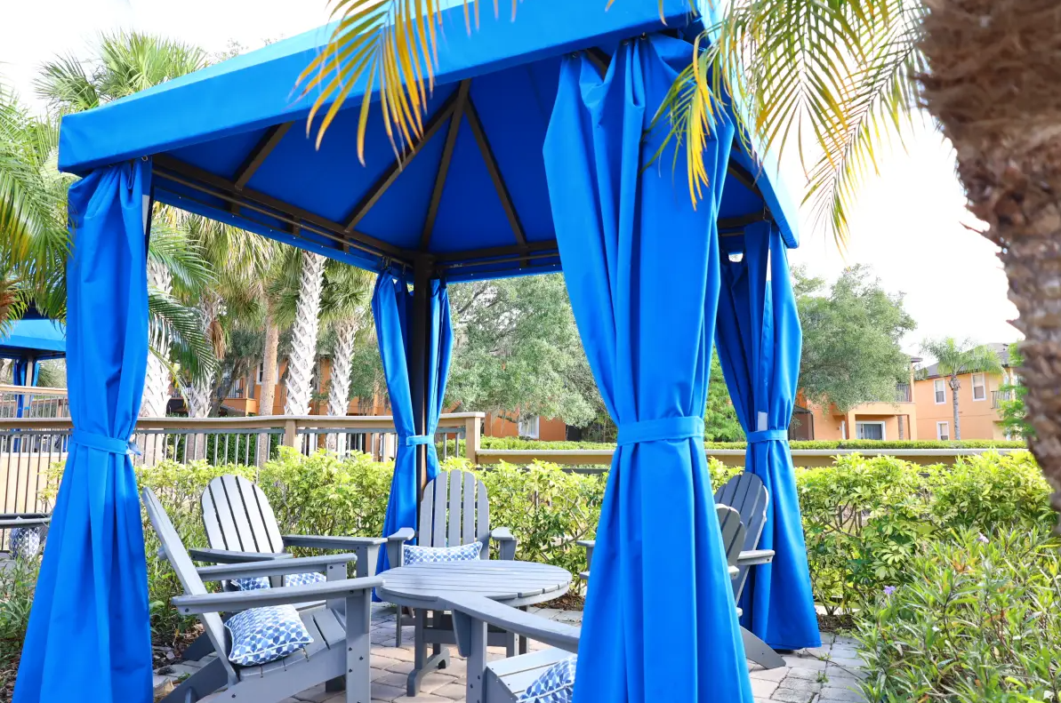 Outdoor seating area with a blue canopy and matching curtains, surrounded by lush green plants and trees, in a residential or resort setting.