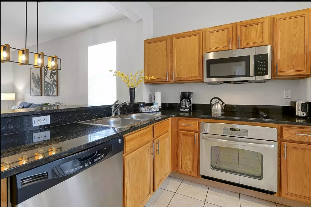 Kitchen with wooden cabinets, black granite countertops, stainless steel microwave, oven, dishwasher, kettle, coffee maker, toaster, and decorative yellow flowers in a black vase.