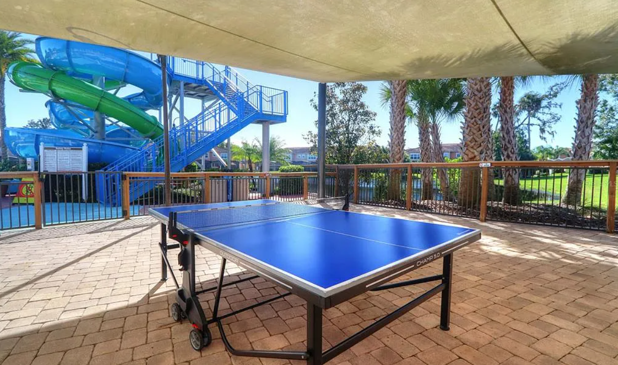 Outdoor recreation area featuring a blue ping pong table under a shaded cover, with a colorful water slide and palm trees in the background.