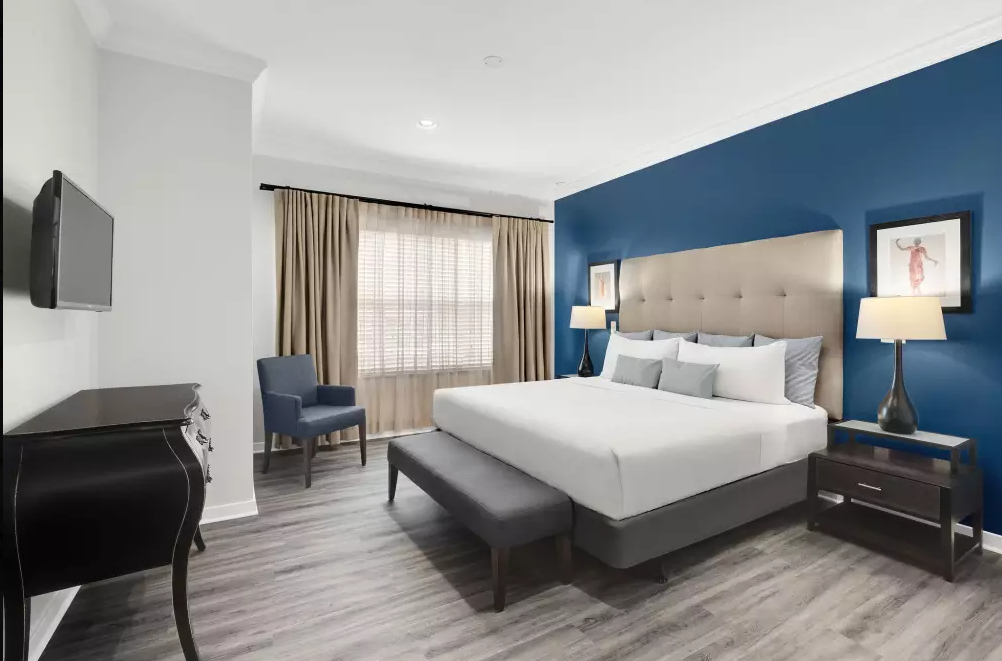 Modern hotel room with a large bed, blue accent wall, bedside lamps, framed artwork, a window with curtains, an armchair, a television mounted on the wall, and a small desk.
