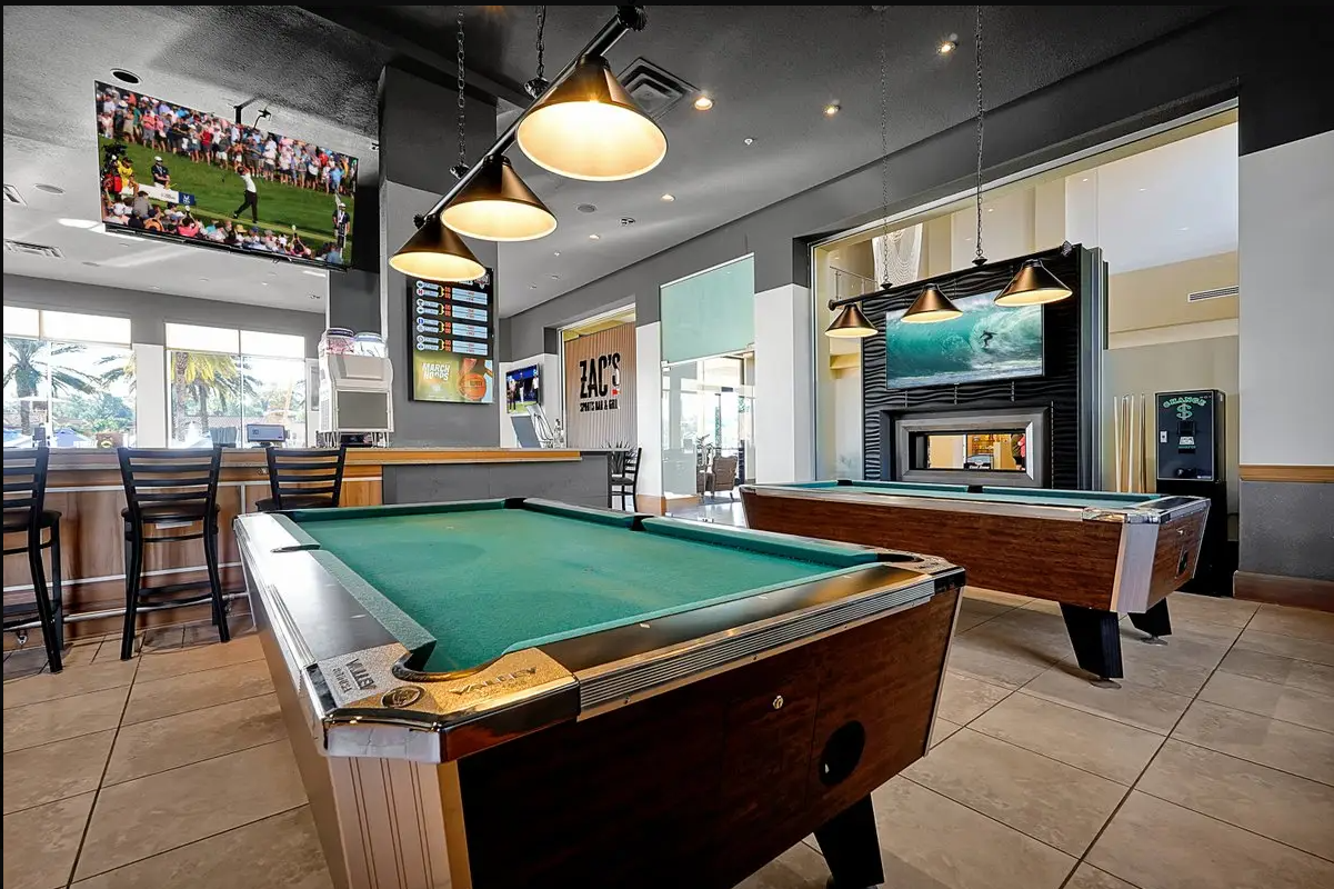 Pool tables in a game room area of a sports bar or restaurant. In the background, a television showing a golf game, a bar counter with chairs, and a large window with palm trees outside.