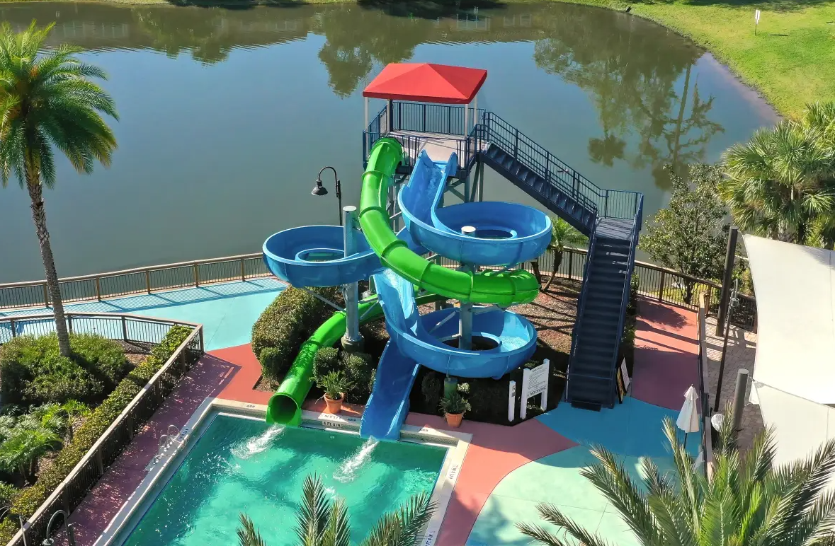 Water park with multiple waterslides, a small pool, a fenced-in area, surrounded by palm trees and a lake in the background.