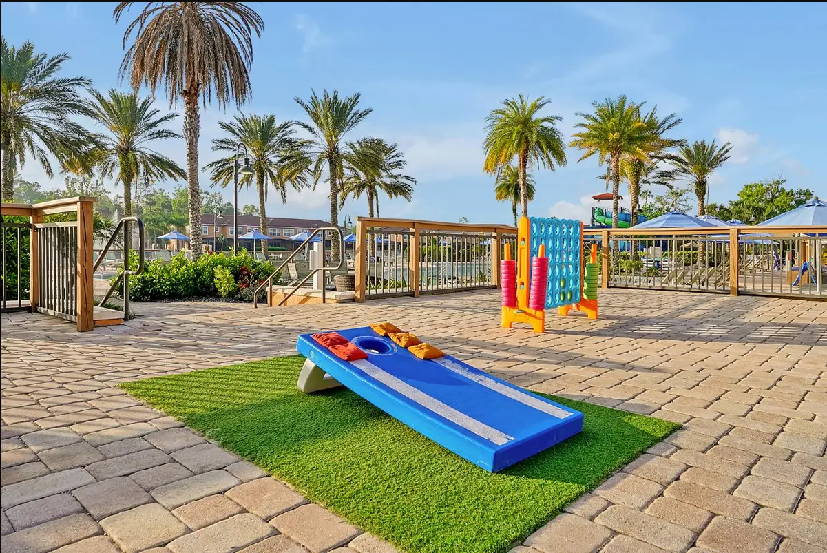 A small outdoor playground with a blue ramp and colorful equipment, surrounded by a sandy brick walkway, palm trees, and umbrellas in the background.