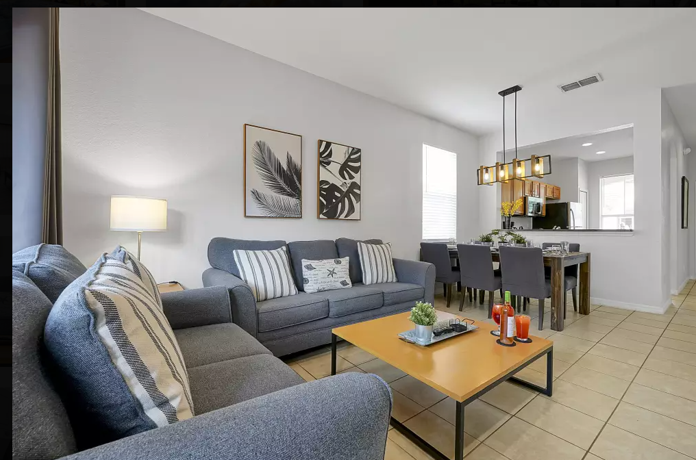 Living room with gray sofas, a wooden coffee table, pillows, and artwork of leaves on the wall, adjacent to a dining area with a dining table and chairs.