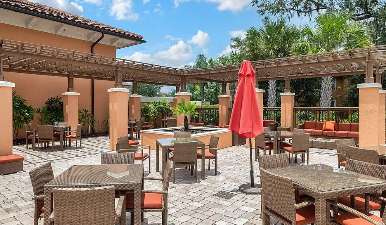 Outdoor patio with wicker tables and chairs, a central fountain with a palm tree, a red umbrella, potted plants, and a beige building with a tiled roof, surrounded by palm trees on a sunny day.