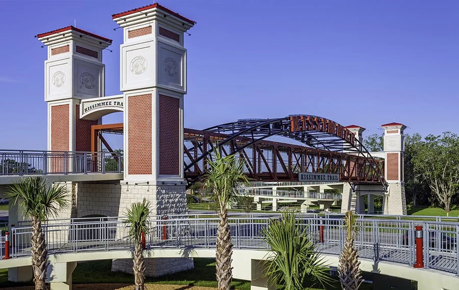 Entrance to Old Town Kissimmee with brick towers, a pedestrian bridge, and palm trees.
