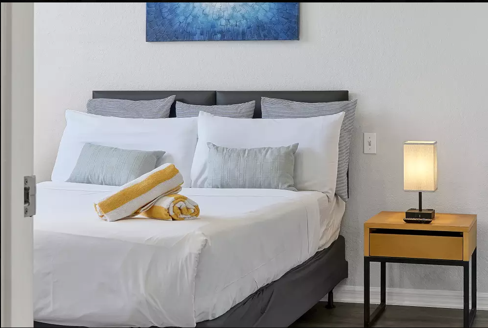 A neatly made bed with white sheets and multiple pillows, including two large striped pillows and two light gray pillows, with rolled towels on top. A wooden nightstand with a small lamp and a telephone is beside the bed. A piece of blue abstract art