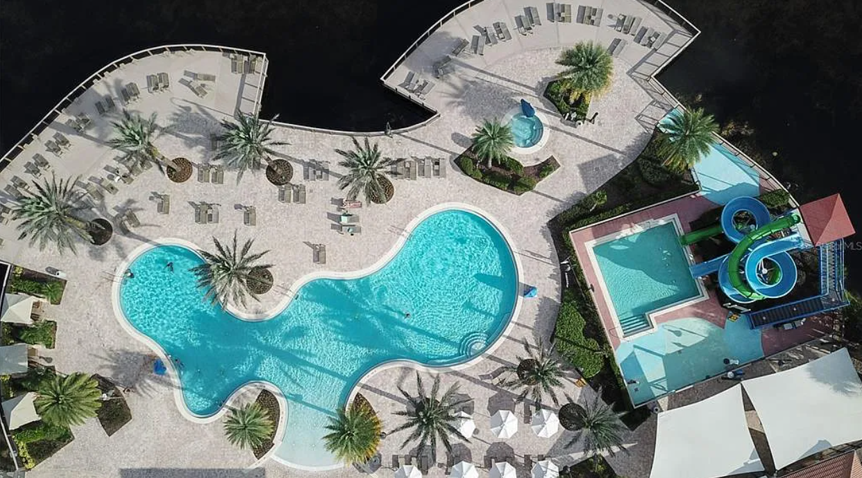 Aerial view of a pool area with a large kidney-shaped pool, a smaller rectangular pool, water slides, and surrounding lounge chairs and palm trees.