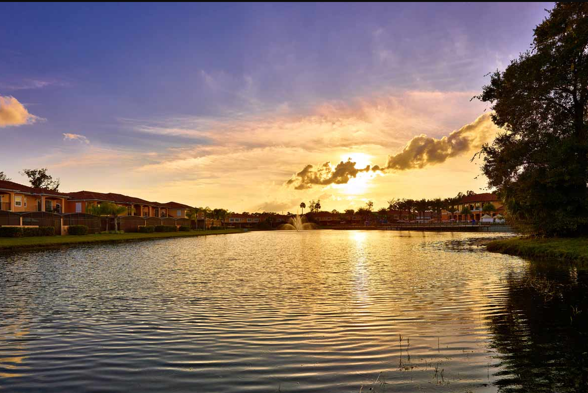 A peaceful lake at sunset with surrounding houses and trees, reflecting the colorful sky.