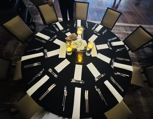 Round banquet table set with white napkins, silverware, and centerpieces consisting of white flowers and candles, in a formal dining setting.