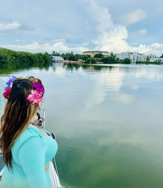 Woman with a flower crown on her head standing on a boat by a calm body of water, with buildings and trees in the background under a partly cloudy sky.