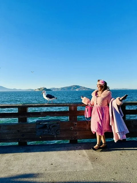 A woman dressed in pink standing on a pier by the water with a bird sitting on the railing nearby, and an island in the background.