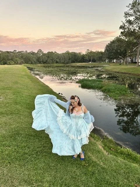 Woman in a blue dress dancing on grass near a pond at sunset.