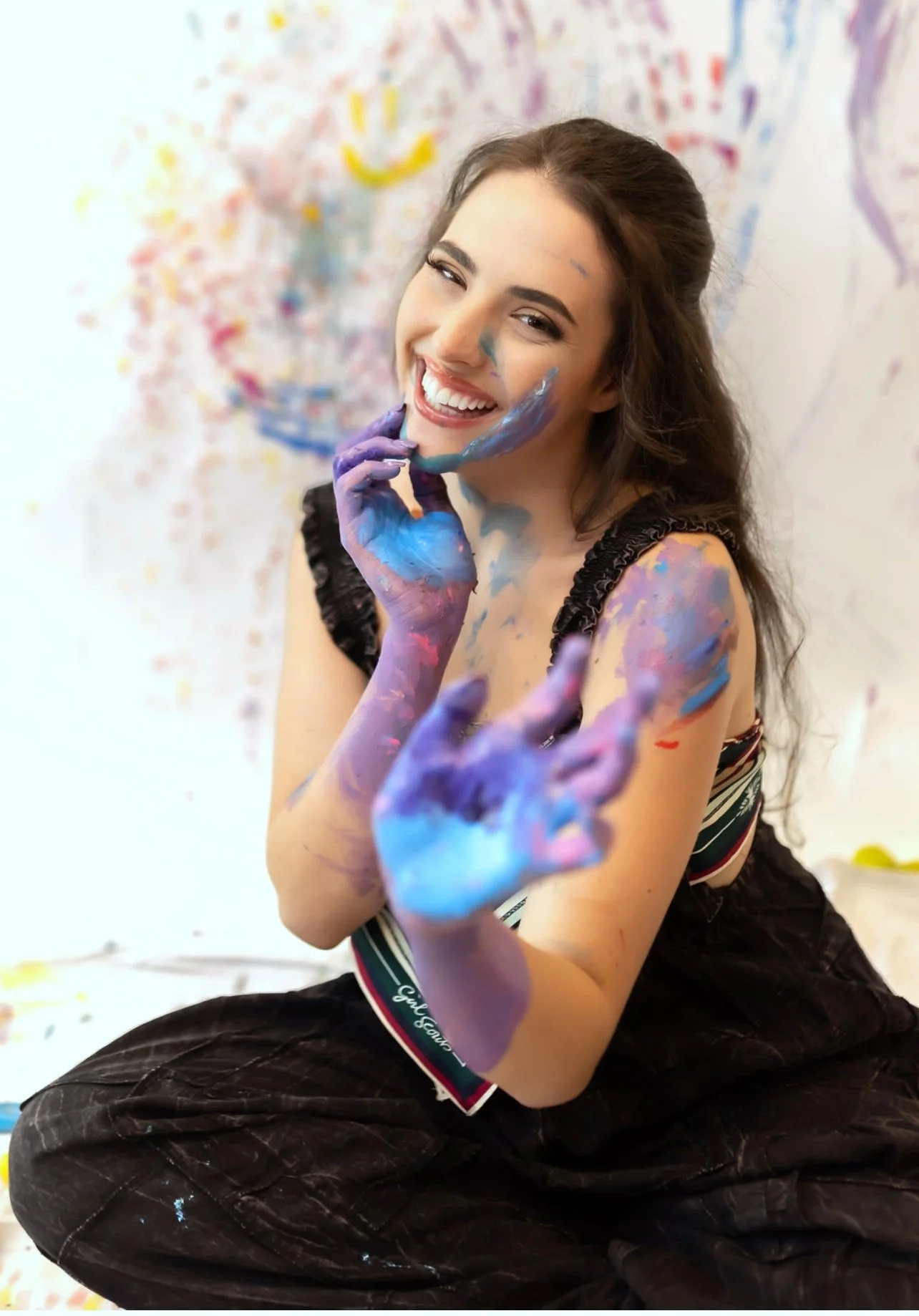 Devyn Wensley of Stained Hands, smiling, covered in colorful paint, sitting on the floor with paint on her hands and face, background decorated with vibrant painted artwork.