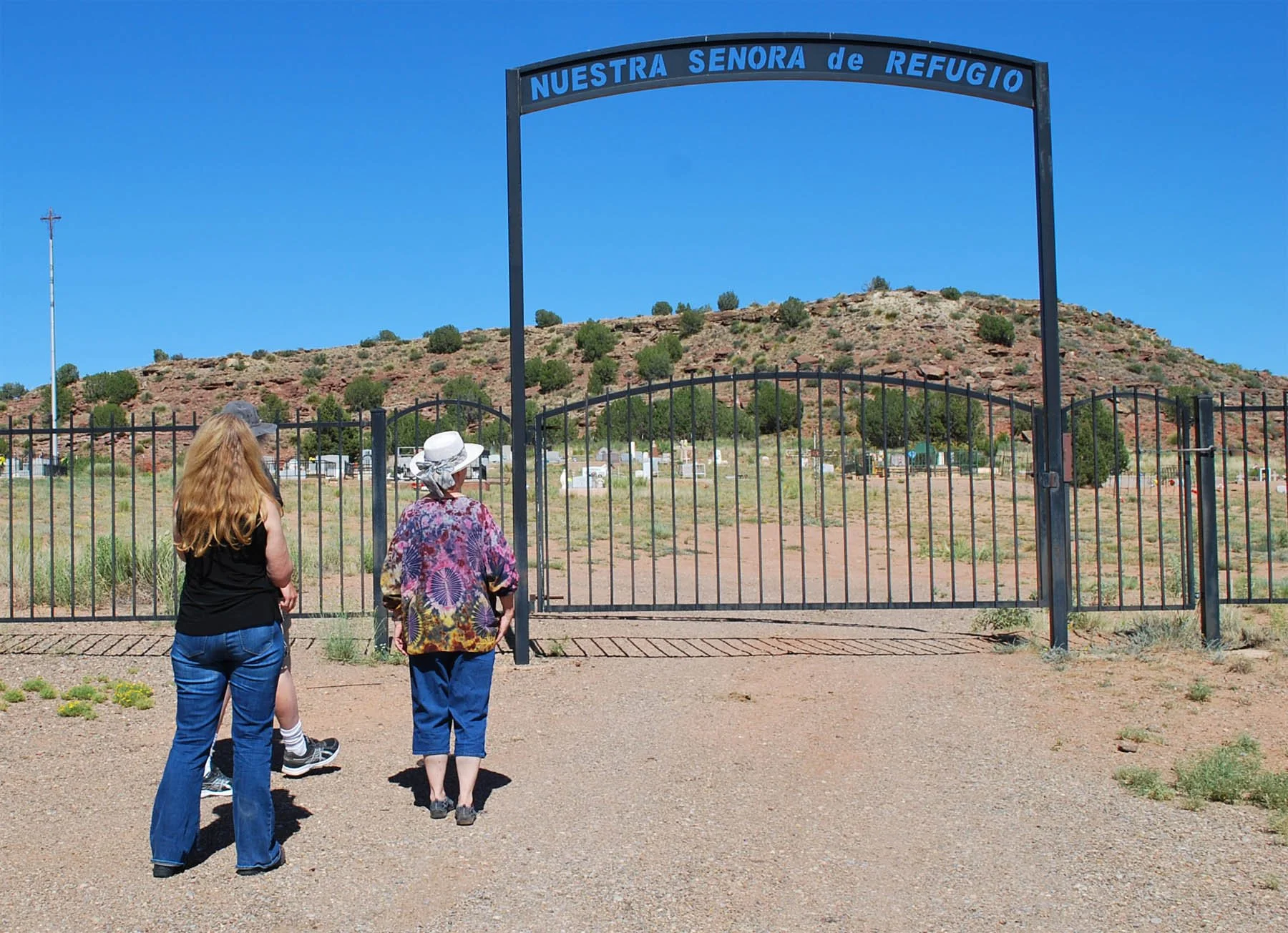 Three women standing outdoors near a black metal fence, looking at a hillside with sparse trees and small graves.
