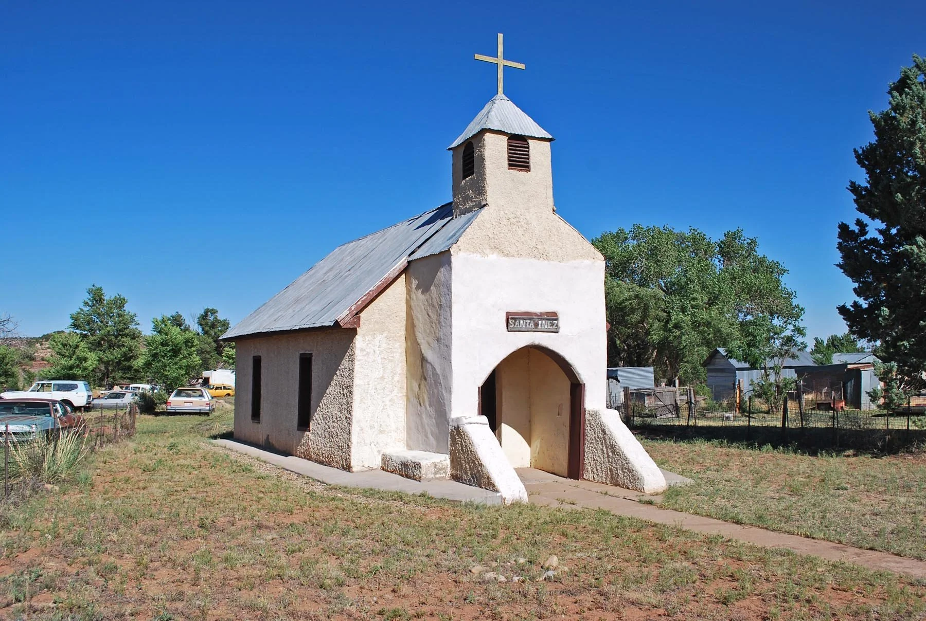 A small white chapel with a steeple topped by a cross, situated in a rural area with multiple parked cars and trees in the background.