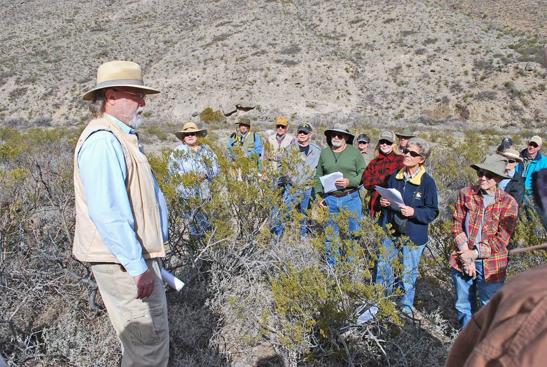 A group of people outdoors in a desert landscape with shrubs, listening to a man in a straw hat and vest who appears to be leading a tour or giving a talk.