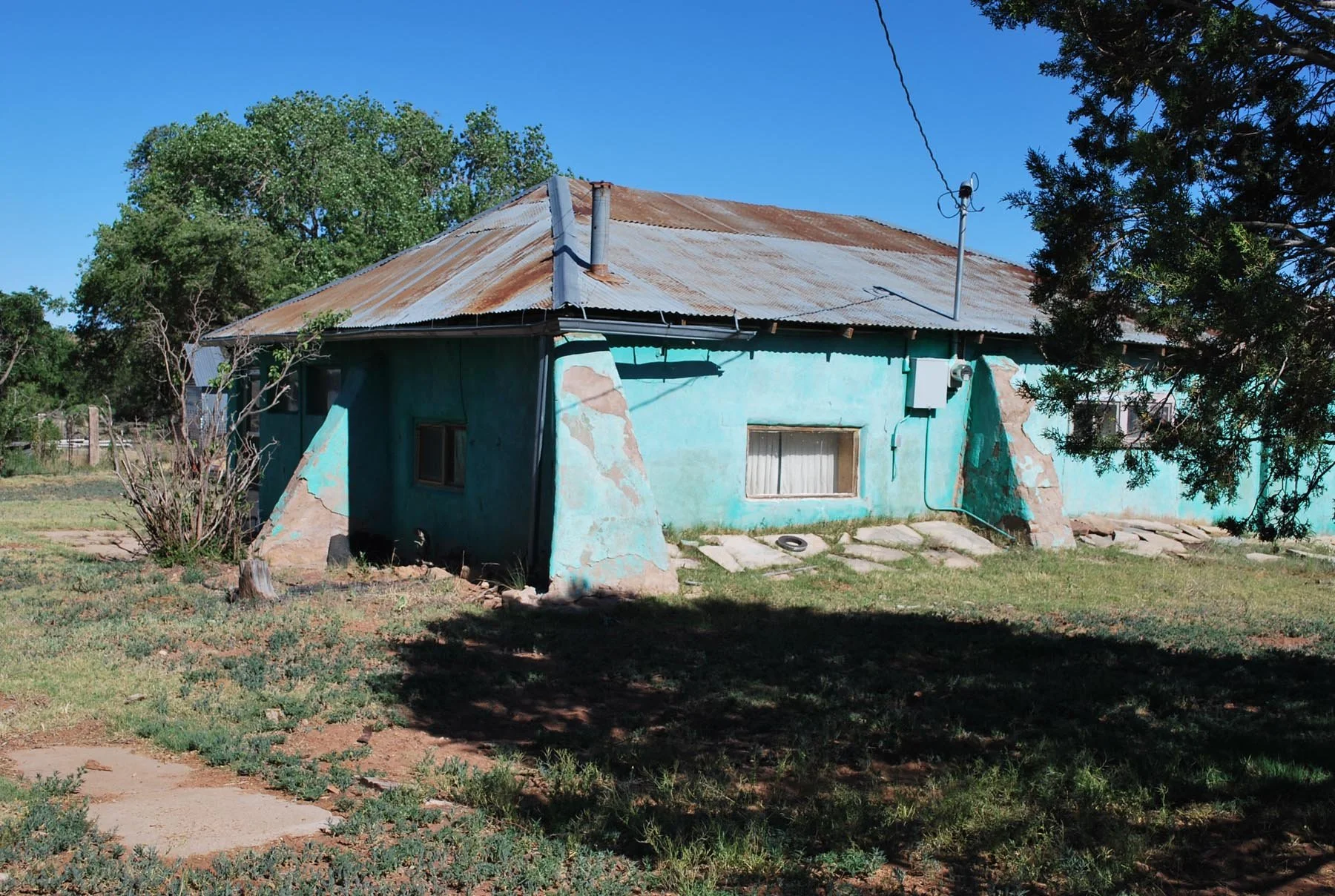 An old, weathered house with a rusted metal roof and peeling turquoise paint, situated in a grassy yard with trees and a fence in the background.